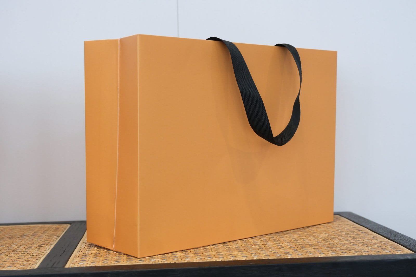 Amber paper bag sample with black flat ribbon handles on a woven table