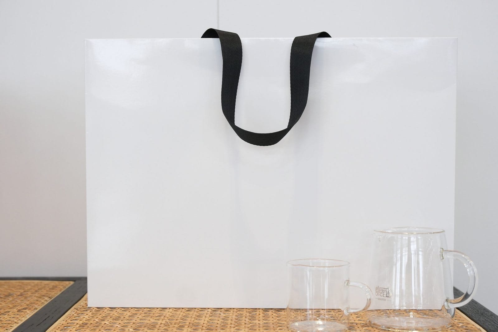 Large white paper bag sample with black flat ribbon handles shown beside glassware for scale