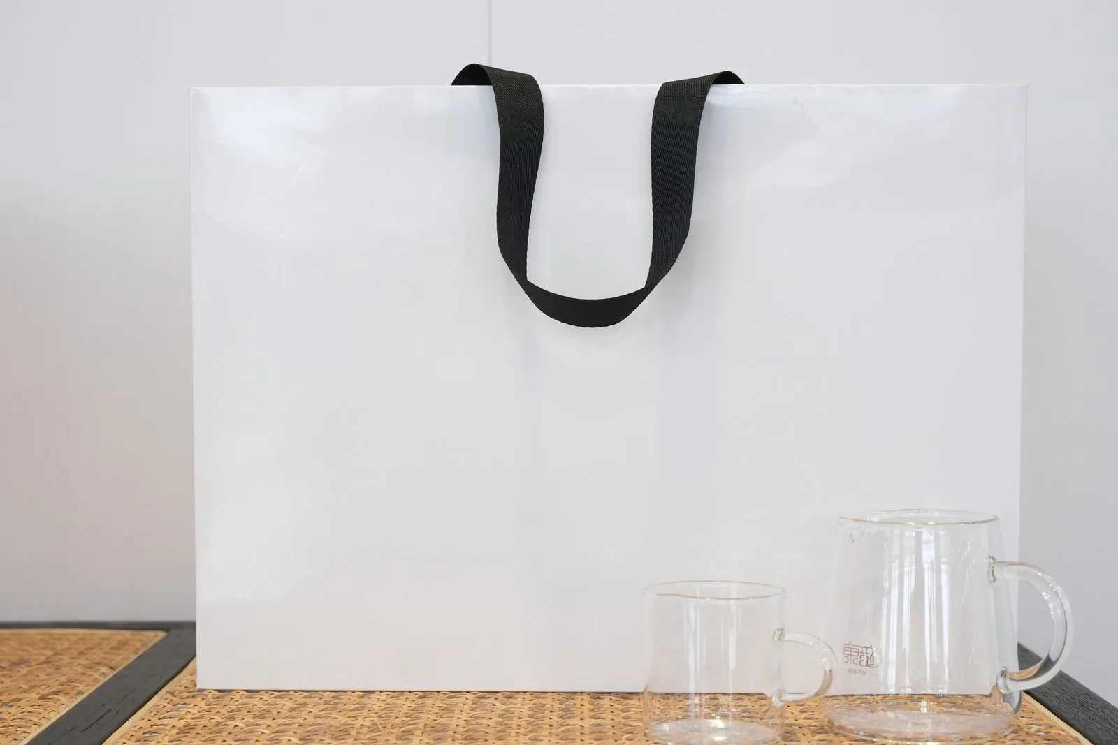 Large white paper bag sample with black flat ribbon handles shown beside glassware for scale